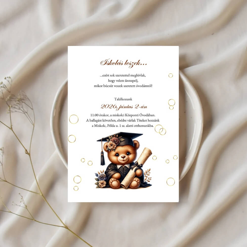 Ovis ballagási dekoráció: diplomás macis meghívó egy elegánsan terített asztalon, fehér tányérra helyezve, száraz virággal díszitve.